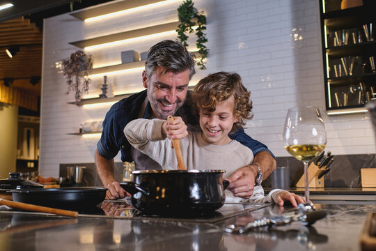 Happy mature man helping smiling boy to stir dish in saucepan while cooking dinner in kitchen at home