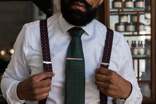 Crop unrecognizable ethnic boss wearing elegant white shirt and suspenders putting on tie lip while preparing for work