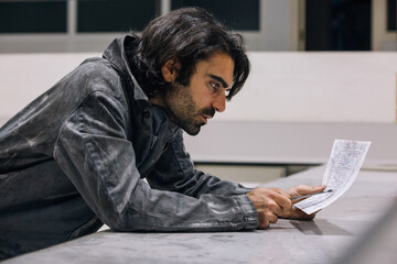 Side view of concentrated bearded adult ethnic male mechanic in workwear reading instruction while working in workshop