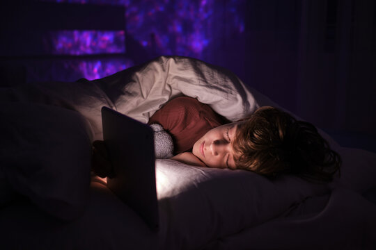 Tired teenage boy sleeping on bed with tablet and toy in dark room with glowing lights