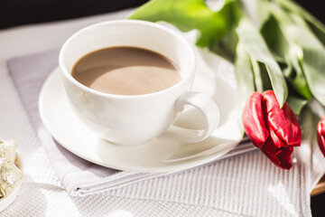 Obraz premium Close-up morning cup of coffee with milk, candy and tulip on the table. Light rays fall on the table