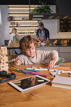 Cheerful boy mixing dominoes near marker pens and book on table against parents cooking lunch in kitchen at home