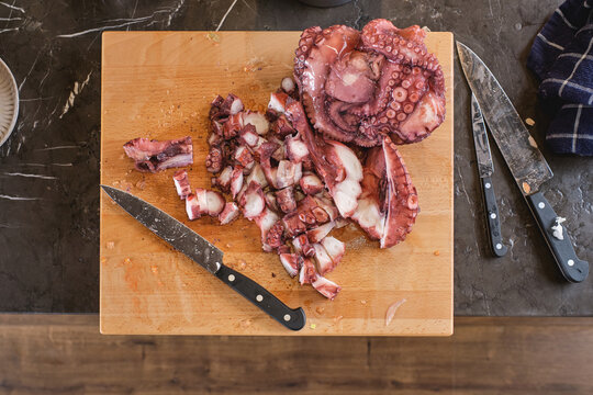 Top view of cut squid tentacles placed on wooden board near knives on table in kitchen at home