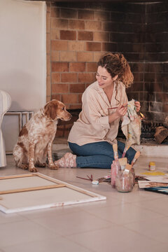 Smiling female artist cleaning paintbrush while sitting on floor in cozy house and looking at fluffy Setter dog