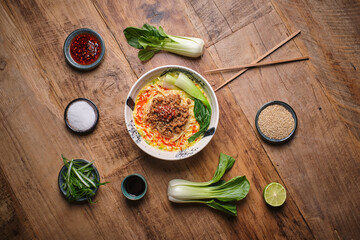 Stock photo of yummy noodles soup with minced meat isolated in wooden background next to sesame seeds and other ingredients.