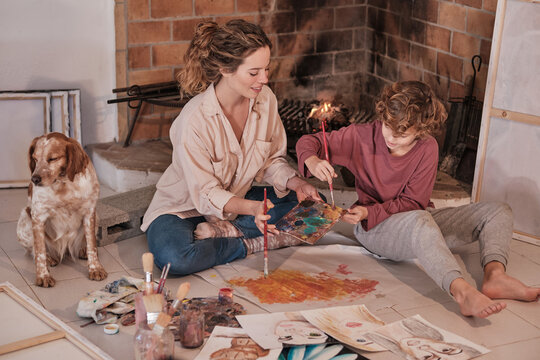 Female artist and teenager sitting on floor with dog and creating painting together while enjoying weekend in house near fireplace
