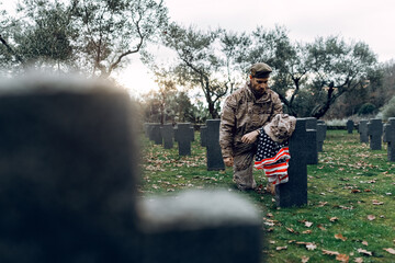 Full body sorrowful soldier in camouflage outfit kneeling down in front of grave in military cemetery on early autumn day