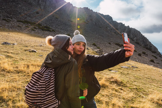 Young content girlfriends with backpacks taking selfie on smartphone standing on grass against mount in sunshine