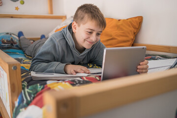 Smiling teenage boy lying on bed and talking to classmates via tablet during online lesson at home