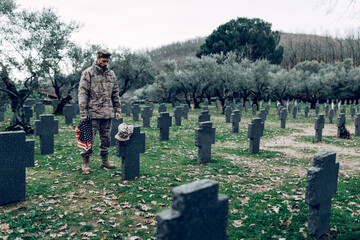 Full body of soldier in uniform standing with American flag while mourning death of warriors at graveyard