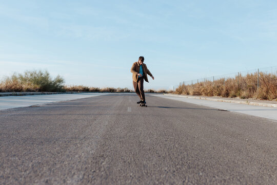 Full body young bearded male skater in stylish wear riding skateboard along asphalt road in countryside