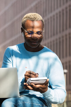 Trendy young African American male freelancer in stylish eyeglasses and casual clothes taking notes in planner while working remotely on laptop on city street