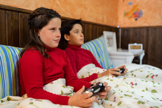 Cheerful teenage brother and sister sitting on bed and playing videogame while using gamepads and enjoying weekend at home