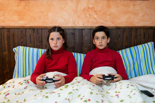 Cheerful teenage brother and sister sitting on bed and playing videogame while using gamepads and enjoying weekend at home