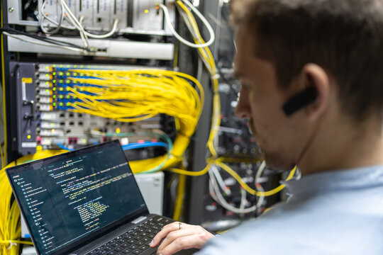 Back view of crop male assistant putting cable in router while using laptop for checking network system