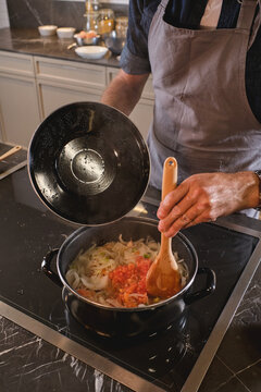 From above unrecognizable man opening lid and stirring chopped onions and tomatoes while preparing sauce on stove at home