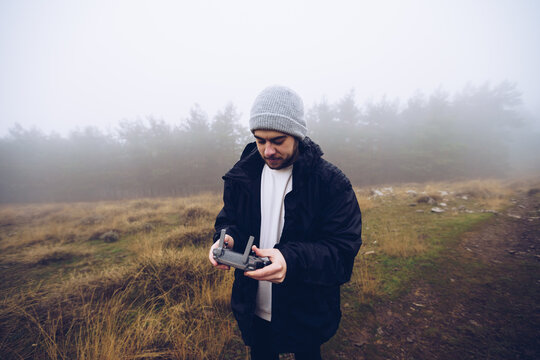 Male operating drone with remote controller while standing on path in foggy field in autumn