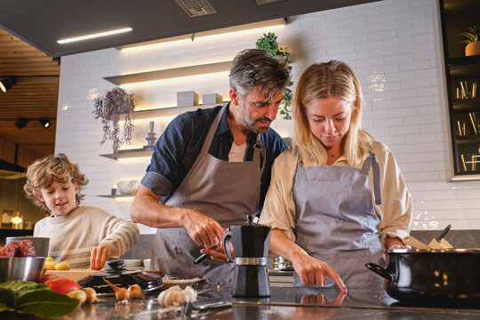 Low angle of mature man and adult woman pushing buttons on modern cooker while preparing food near boy in modern kitchen at home