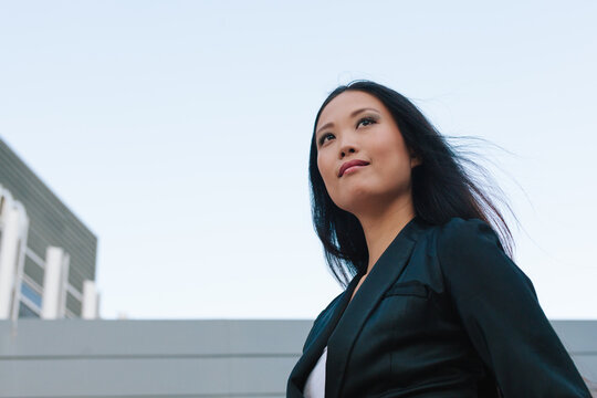 Low Angle Of Determined Well Dressed Asian Female Entrepreneur Standing With Hand In Pocket In City Street And Looking Away