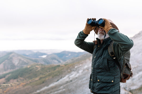 Senior male traveler in outerwear looking through binoculars while standing in highland terrain and admiring picturesque landscape during winter holidays in Caceres