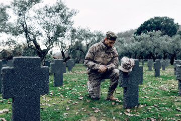 Full body sorrowful soldier in camouflage outfit kneeling down in front of grave in military cemetery on early autumn day