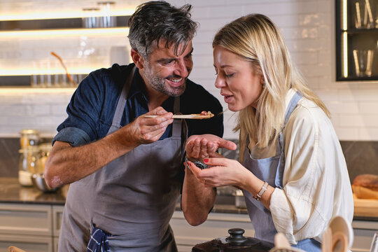 Mature man smiling and sharing dish with adult woman while preparing dinner in kitchen at home