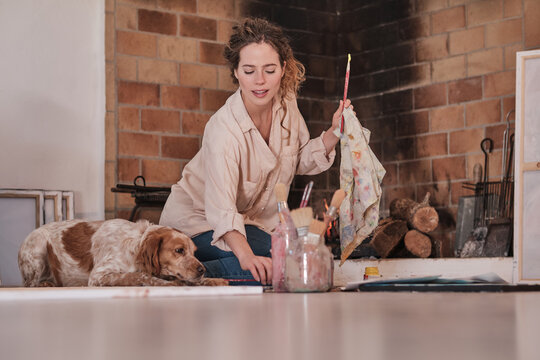 Smiling female artist cleaning paintbrush while sitting on floor in cozy house with fluffy Setter dog