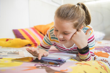 Happy schoolgirl lying on bed with tablet and having video call with classmates during online lesson from home