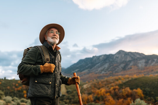 Side view of elderly male wanderer with wooden stick standing on hill and admiring scenic view of highlands in Caceres