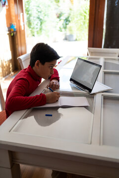 High Angle Side View Of Smart Schoolboy Sitting At Table With Laptop And Writing In Notebook While Doing Homework Alone