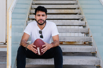 Confident young bearded ethnic male in white t shirt sitting on stairs on street and looking at camera with rugby ball in hands
