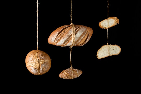 Loafs and pieces of fresh artisan crispy bread of various shapes and sizes hanging on ropes against black background