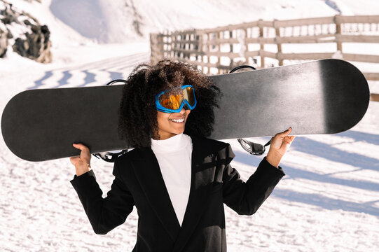 Happy ethnic sportswoman in stylish wear and protective glasses carrying snowboard on snowy mount in sunlight