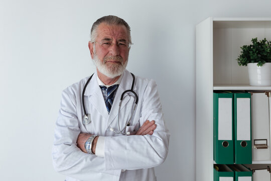 Senior male doctor with gray beard and folded arms in robe with stethoscope looking at camera in clinic
