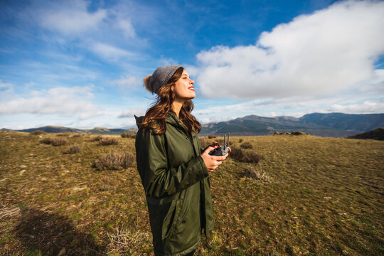 Young female tourist with pouting lips controlling UAV with remote controller while looking away on lawn in Europe
