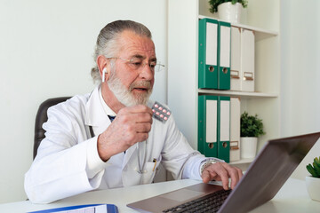 Elderly male doctor in earbud showing medications while talking on video chat against netbook during online health consultation in hospital