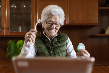 Elderly woman with gray hair in wireless earbuds with toothbrush and floss against tablet during online health consultation in house