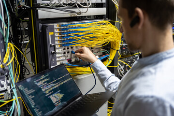 Back view of crop male assistant putting cable in router while using laptop for checking network system
