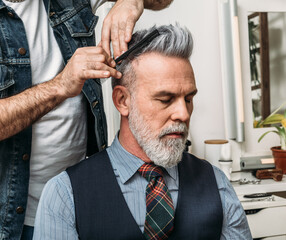 Crop anonymous barber styling hair of elegant well dress bearded middle aged male customer sitting on chair and looking away in modern studio