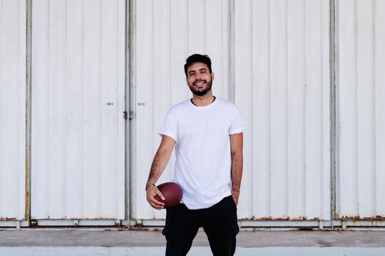 Self assured young bearded ethnic male in white t shirt holding pigskin while playing American football on sunny day