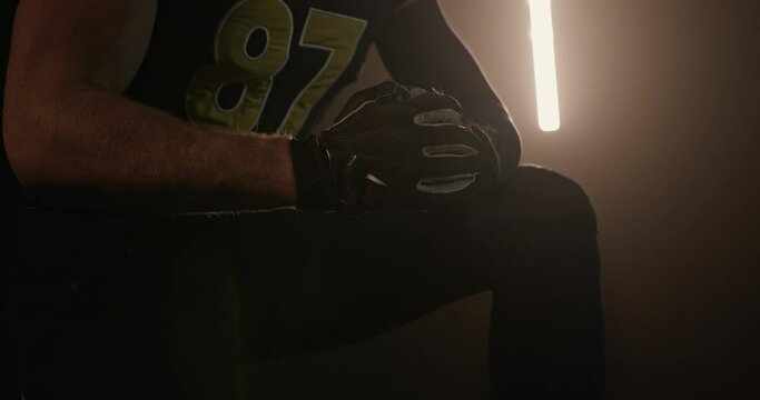 Crop rugby player cracking knuckles in dark studio. Crop unrecognizable male American football player in uniform and gloves sitting in dark studio and cracking knuckles