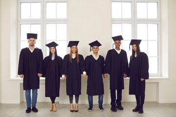 General portrait of a group of happy and successful multiracial university graduates in academic dresses standing holding hands. Education, graduation and people concept.