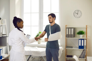 African american woman doctor therapist greeting caucasian man patient with handshake at hospital ward. Professional consultation and injured hand medical treatment. Medicine and health insurance