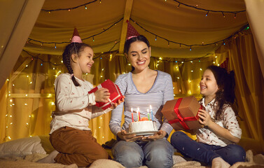 Fototapeta na wymiar Mom gives birthday gifts to her twin daughters and a cake sitting in a cozy bed tent. Family in holiday hats celebrates at home in a tent bed decorated with yellow LED fairy lights.