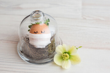 Fototapeta na wymiar Cute egg with eyes, a flower wreath, in a medical mask celebrates Easter alone, in self-isolation under a glass cover, beautiful spring flowers