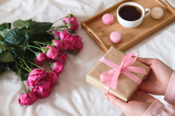 Woman's hand holding a gift. Cozy holiday morning composition with roses and cup of coffee with macaroons on wooden tray