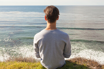 Stress-free wellness. Relaxed young man alone sitting on top a cliff facing the ocean view. 
