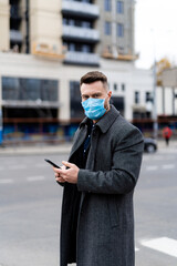 Stylish man in coat and mask standing near modern buildings and holding phone. Coronovirus pandemic. World disease.