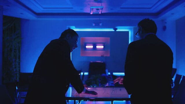 Two men in black suits meeting at dark office room and using a modern, futuristic big touch screen tablet. Businessmen looking at map illuminated on digital table, discussing and talking . Slow Motion