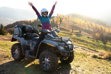 Happy woman driver in protective helmet enjoying extreme riding on ATV quad motorbike in summer mountains at sunset. - obrazy, fototapety, plakaty